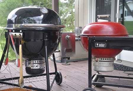 Large black charcoal kettle gril with tools hanging down next to a medium size red charcoal kettle on an outdoor deck.