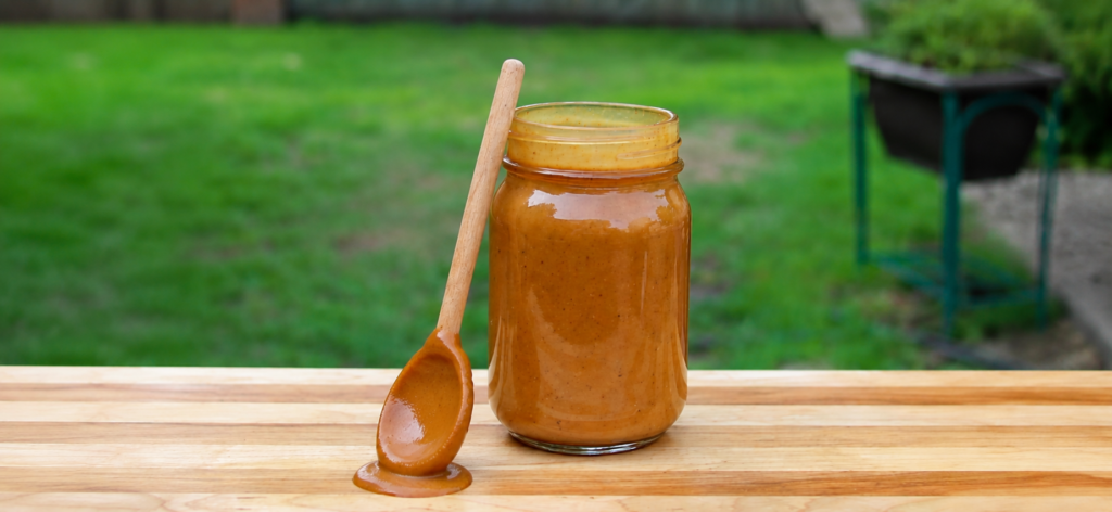 Mustard BBQ sauce in a clear jar with a spoon resting against the jar