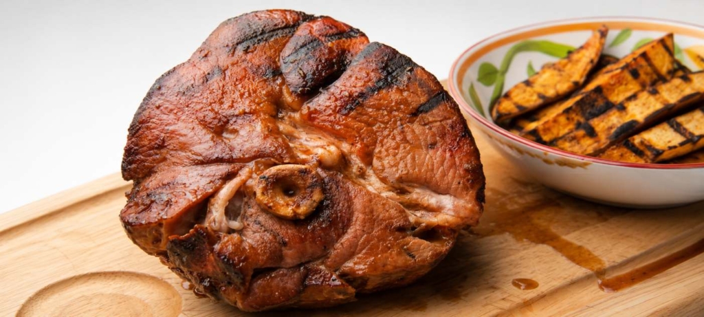 Double smoked ham on a cutting board with grilled sweet potato wedges in a bowl