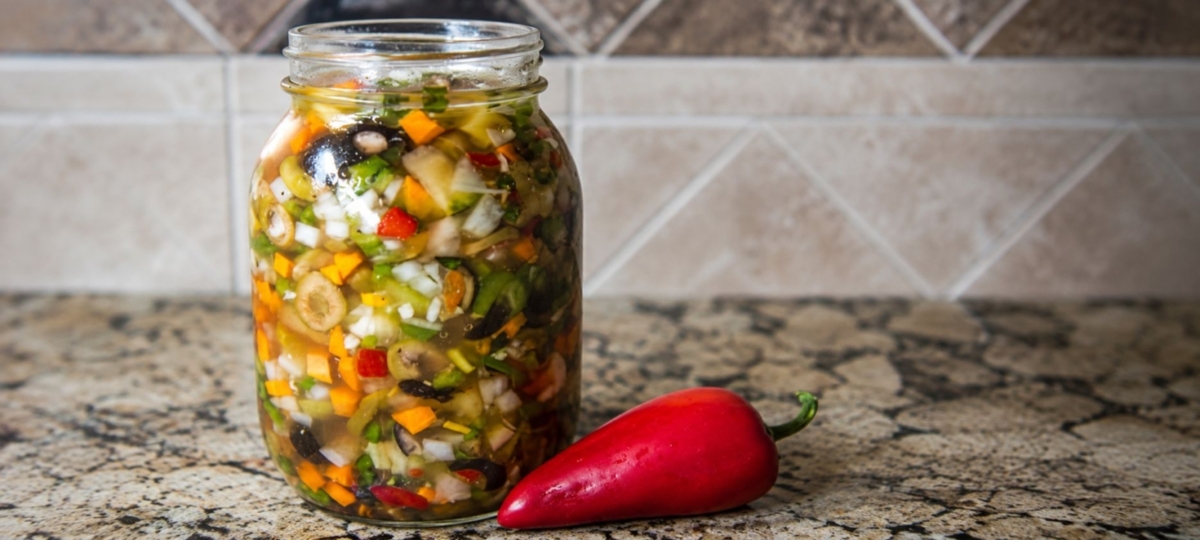 Glass jar of hot giardiniera on a marble countertop.