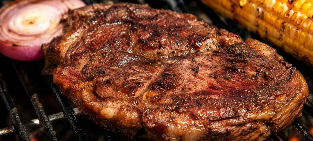 Perfectly seared chuck steak on a grill with corn and grilled onion.