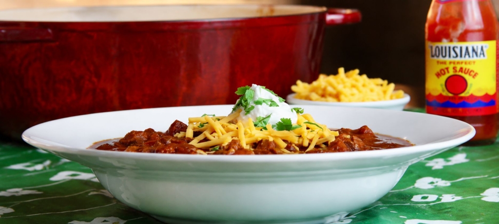 Chili con carne in a bowl with cheese and sour cream with a bottle of hot sauce and a bowl of cheese