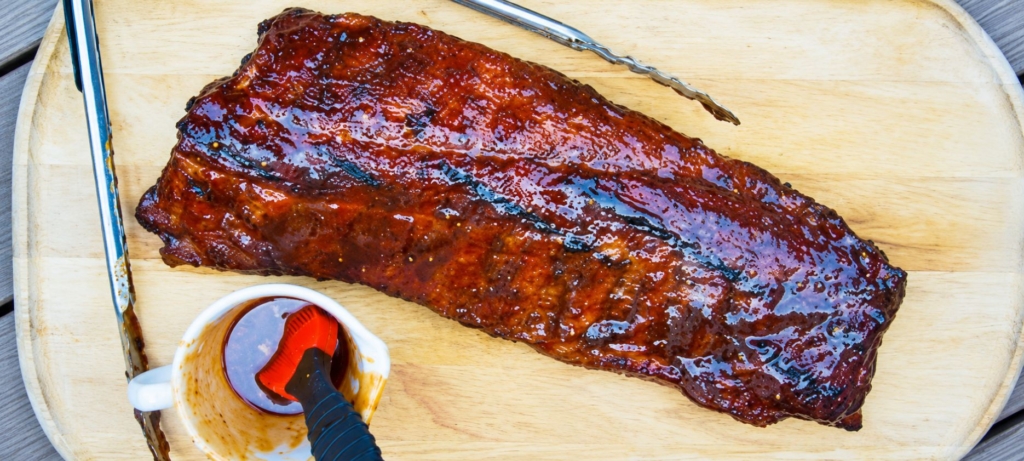 Rack of ribs, tongs, and cup of BBQ sauce on a cutting board.