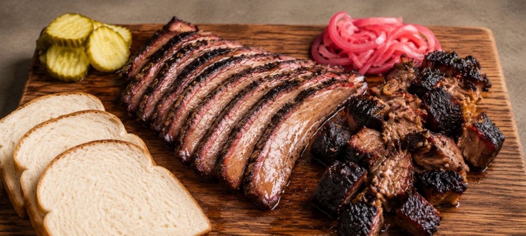 Sliced smoked brisket with burnt ends, white bread, pickle chips, and pickled red onions arranged on a wooden cutting board.