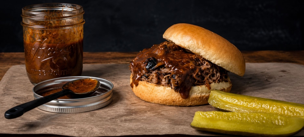 Jar of mustard sauce, a pulled pork sandwich with mustard sauce and two pickles