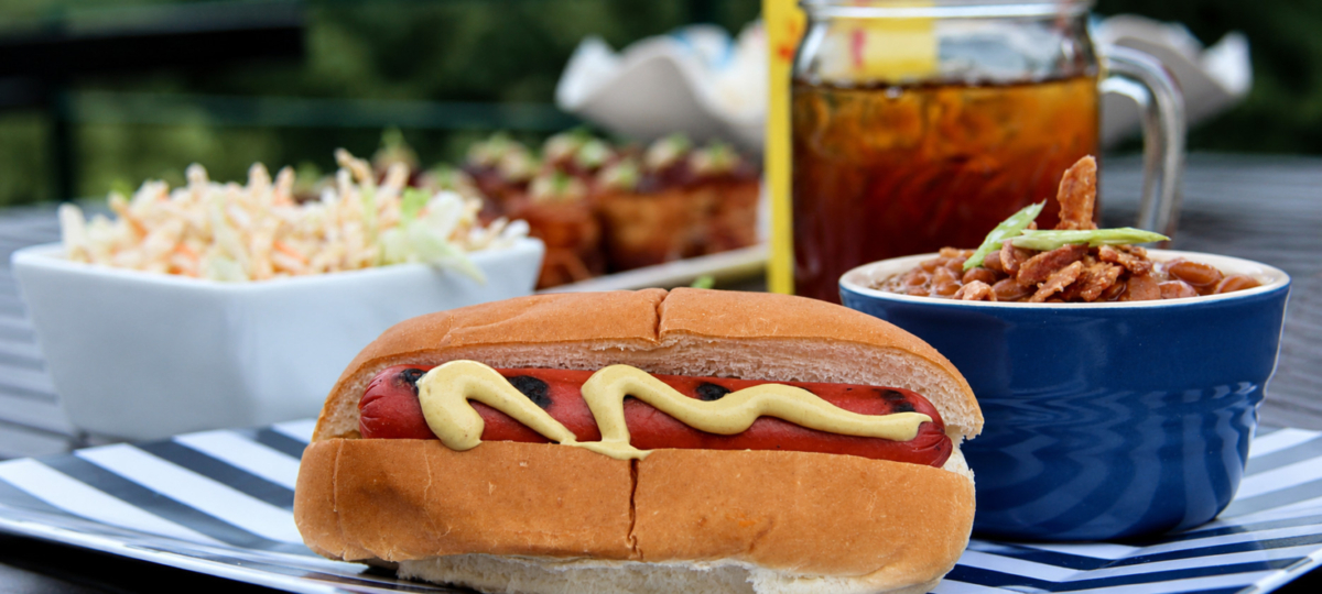 Hog dog topped with mustard in a hot dog bun alogside coleslaw, beans, and tea