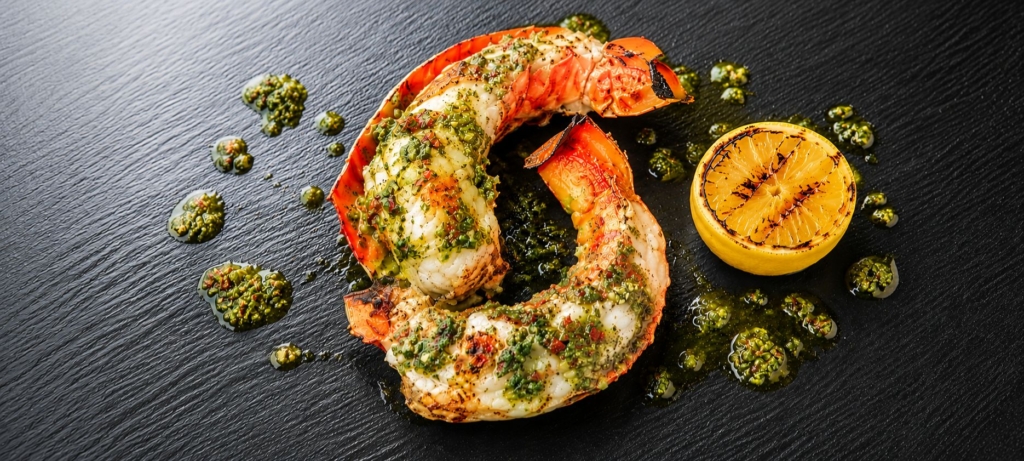 Grilled split lobster tail topped with Chimichurri sauce alongside a grilled lemon half.