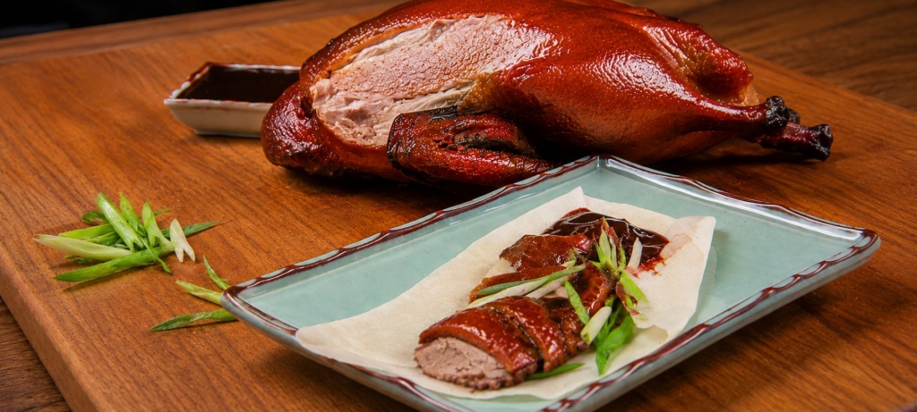 Smoked Peking duck slices on a pancake with sliced scallions and hoisin sauce on a green plate and a whole duck in the background
