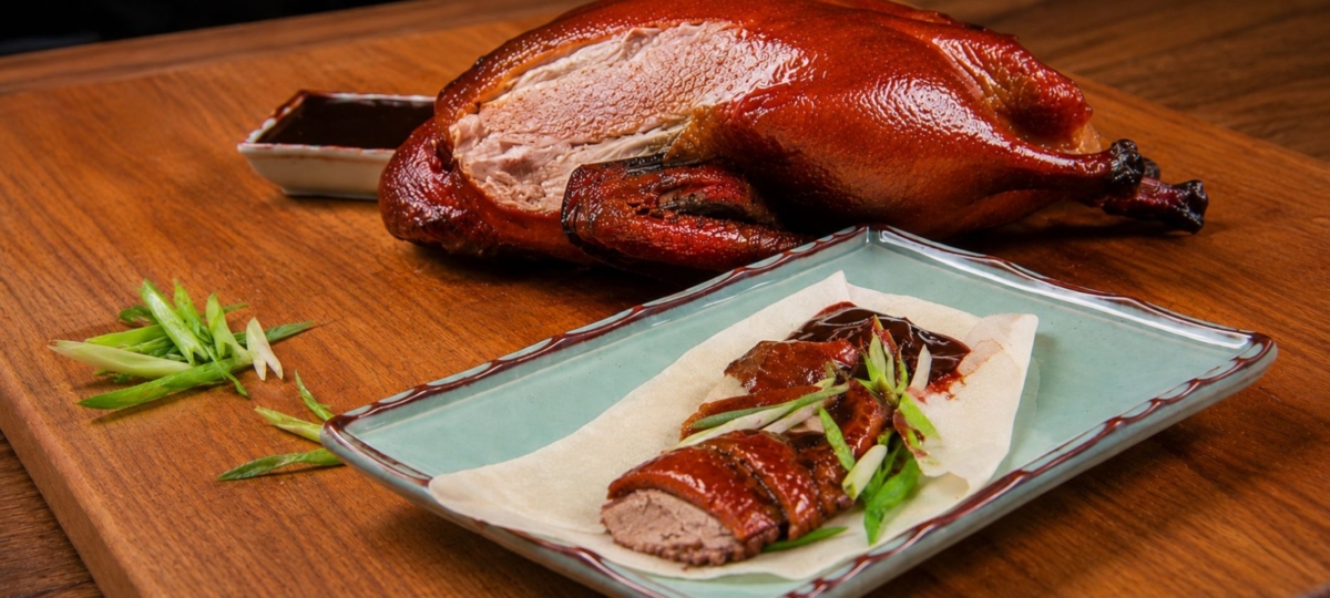 Smoked Peking duck slices on a pancake with sliced scallions and hoisin sauce on a green plate and a whole duck in the background