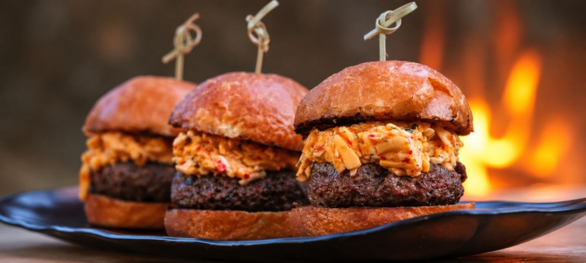 Three grilled pimento cheese burger sliders on toasted brioche buns with creamy pimento cheese and flames softly blurred in the background.