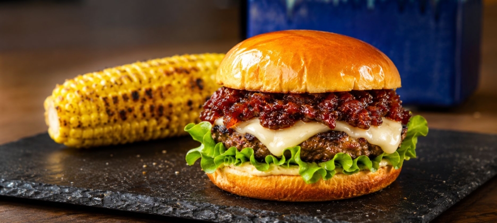 Burger topped with cheese and bacon jam with an ear of corn in the background.