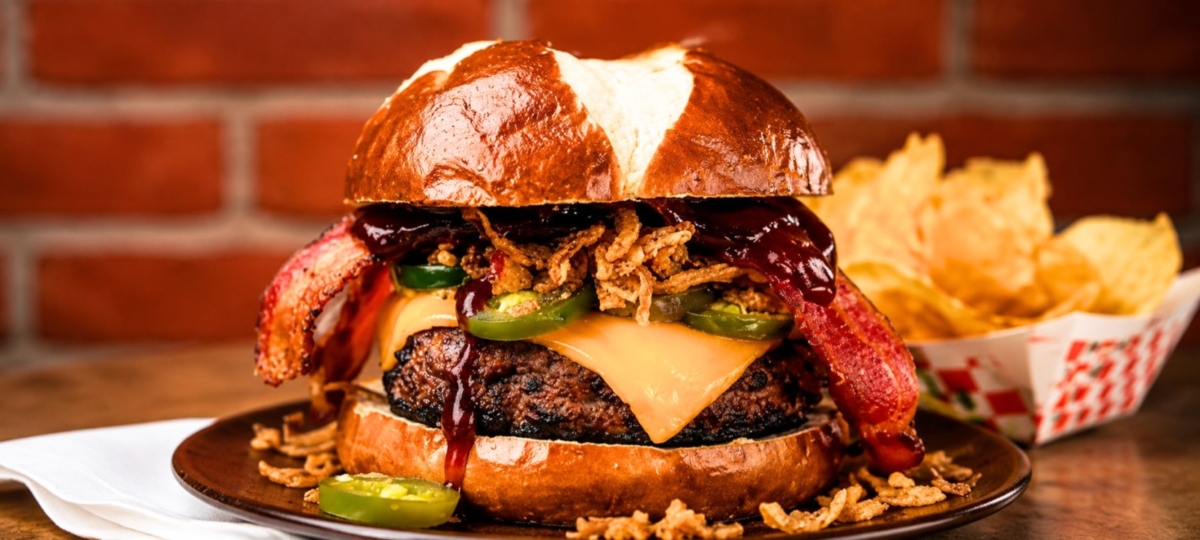 BBQ bacon cheeseburger with crispy fried onions, jalapeños, and cheddar on a pretzel bun, served with potato chips.
