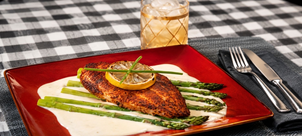Grilled catfish sitting on asparagus and sauce, topped with a grilled lemon. Served with