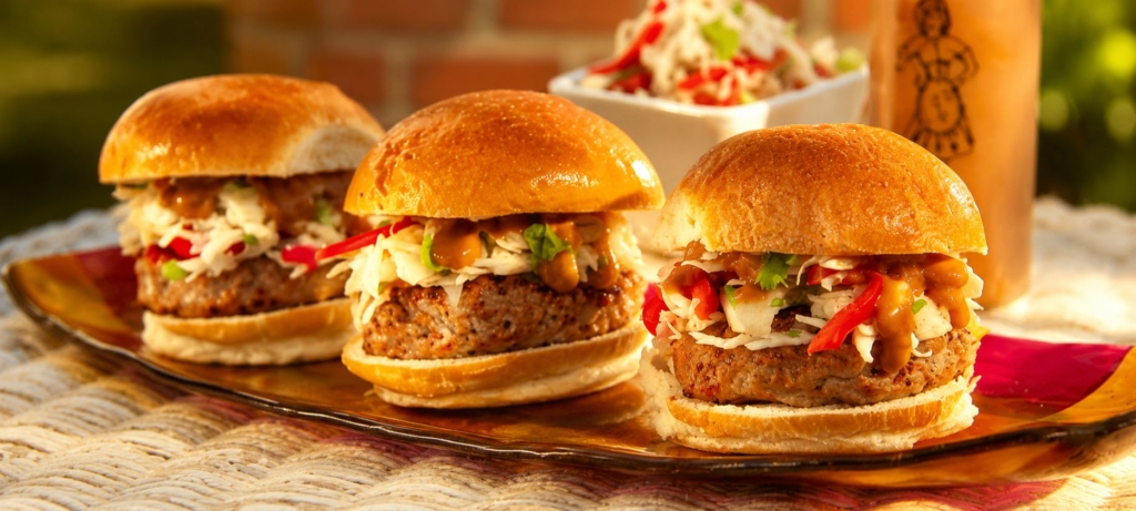 Three pork satay sliders topped with slaw and peanut sauce on a platter with more slaw and a bottle of peanut sauce in the background.