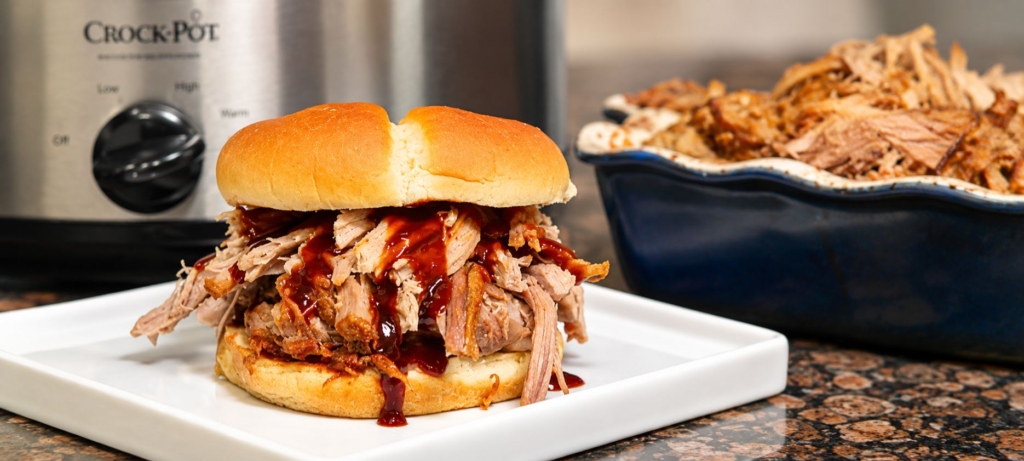 Crockpot pulled pork sandwich on white plate with a Crock Pot and a bowl of pulled pork in the background