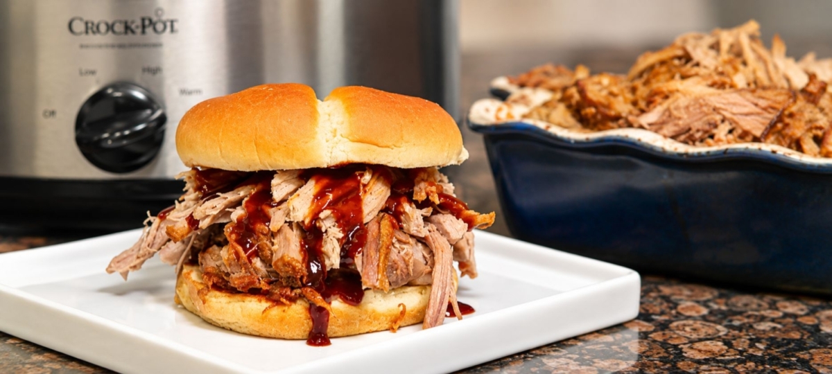 Crockpot pulled pork sandwich on white plate with a Crock Pot and a bowl of pulled pork in the background