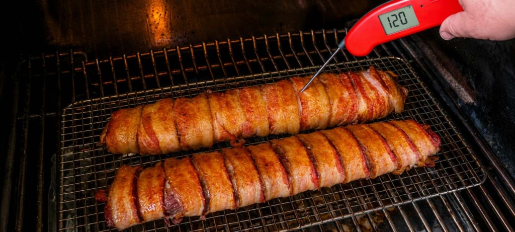 bacon wrapped venison backstrap on grill and a red thermometer