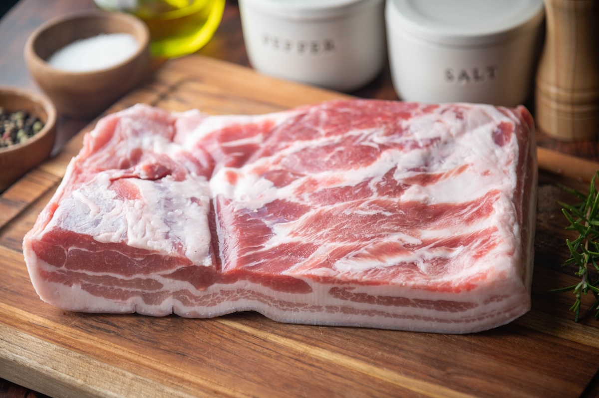 raw slab of pork belly on a wooden cutting board with seasonings in the background.