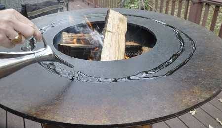 Large round firebowl filled with burning wood. A hand is pouring oil on the flat perimeter surface.