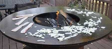 A large round firebowl with a wide griddle surface circling the hot wood fire. Bacon and chopped onions are all around the griddle.