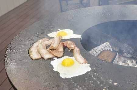 A large round fire bowl with a flat griddlw wrapped around the wood fire. Bacon and eggs are cooking on the griddle.