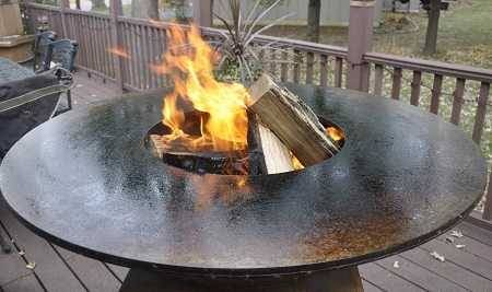 A large round fire bowl with a blazing wood fire in the center. It is on an outdoor deck.