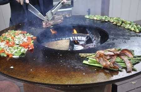 A large round fire bowl with a wide griddle surface circling the hot wood fire. Meats and vegetables are cooking all around the griddle. A man is cutting into a steak with a large knife.