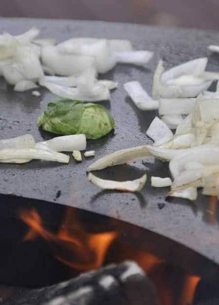 Curved metal surface with a fire underneath. Chopped onions and Brussels sprouts are cooking on the surface.