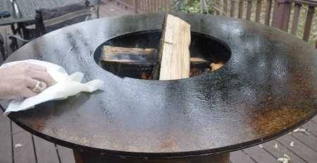 Large round firebowl filled with burning wood. A hand is wiping down the flat perimeter surface with a paper towel.