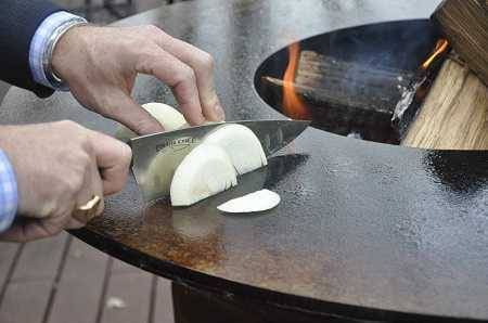 A large round firebowl with wood buring in the center. A man is cutting onions right on the flat metal surface surrounding the fire.