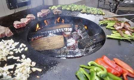 A large round fire bowl with a wide griddle surface circling the hot wood fire. Meats and vegetables are cooking all around the griddle.