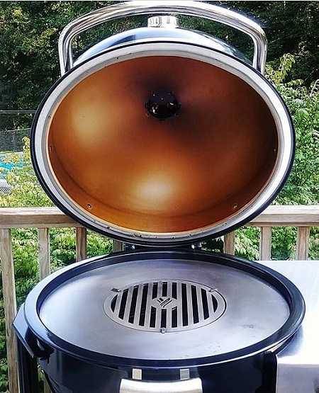 Kamado with dome open to show heavy metal round griddle inside. On an outdoor deck with green bushes in the background.