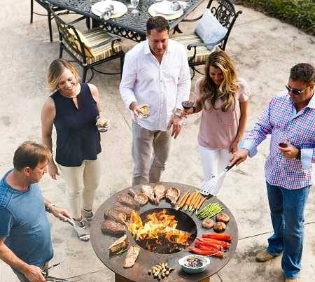 Men and women standing around a large round fire bowl with a griddle cooking surface wrapped around the blazing wood fire. They are cooking meat and vegetables and drinking wine.