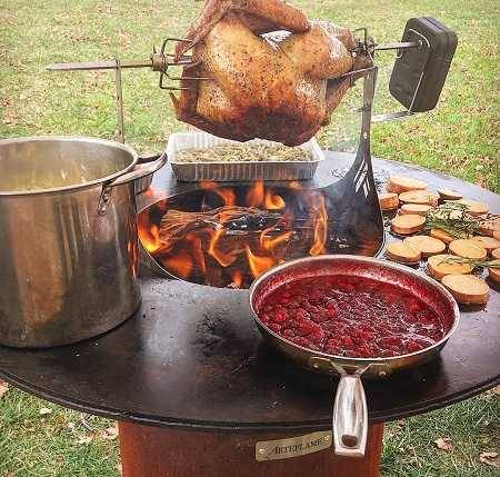 A large round fire bowl with a flat griddle surface wrapped around the central wood fire. Pots and pans filled with food are cooking on the griddle and a chicken on motorized rotisserie is hovering over the fire.