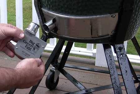 Two hands on the left hold a shiny steel box labeled "Big Green Egg". They appear ready to attach the box to a pipe at the bottom of a green kamado. There is a while fence and green grass in the background.