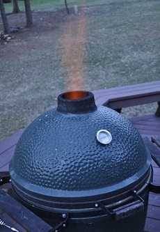 A green kamado on an outdoor deck with grass in the background. Flames hoot out the top.