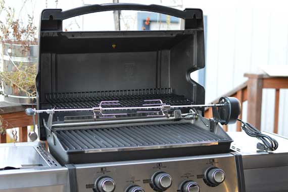 A gas grill with the lid up showing a rotisserie spit in place.
