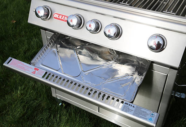 Shiny stainless steel gas grill with a foil covered tray slide out from under the control knobs.