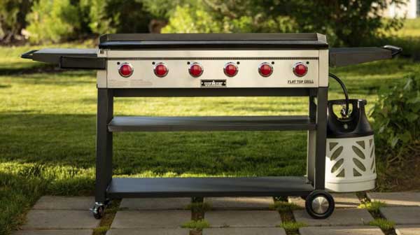 Large outdoor griddle on four legged cart placed on a brick walk way. Six red dials are on the front.