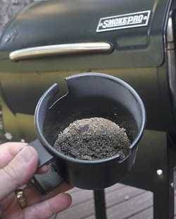 A hand holding a cup of ashes with a pellet smoker in the back ground.