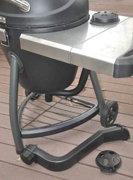 A black oblong structure on three legs with a triangular shelf at the bottom on an outdoor deck. Some black parts are laying on the deck.