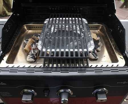 A gas grill from above with the lid up showing a metal tray inside. An aluminum pan with a cooking grate on top is in the center of the tray. Wood and charcoal burns on each side of the pan.