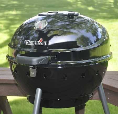 Black charcoal kettle grill on an outdoor deck with grass in the background. A Char-Broil logo is attached to the hood. There are one inch diameter holes circling the lower section of the kettle.