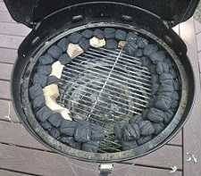Black charcoal kettle grill from above. The lid is up showing charcoal arranged around the edge of the lower charcoal grate in a C shape with chunks of wood mixed with the charcoal.