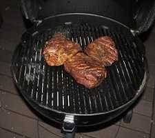 lack charcoal kettle grill at night with lid up showing three smokey hunks of pork on the cooking grate.