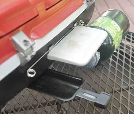 A red and black metal object place on a metal grate. A black metal tray is pulled out on the lower left. A small green canister is attached on the right.