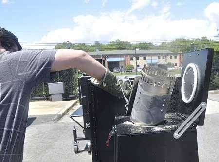 A man dumps hot charcoal into a large black smoker box attached to a larger smoker box. He is in a parking lot with a wire fence, houses and blue sky in the background.