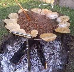 A metal disc placed above a wood fire, loaded with meat in sauce with  tortillas all around the edge. Outdoors on grass.