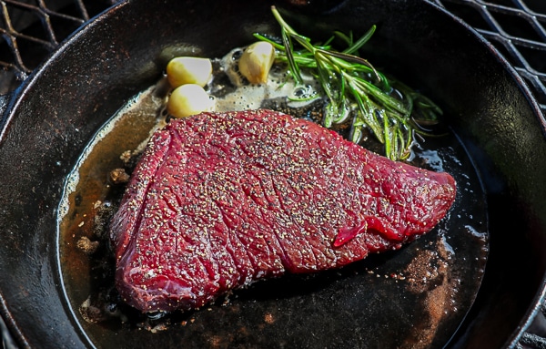 Elk steak being cooked in a cast iron skillet along with fresh rosemary and garlic cloves