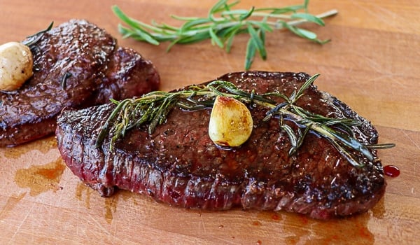 Cooked elk steak on a cutting board with fresh rosemary and garlic clove